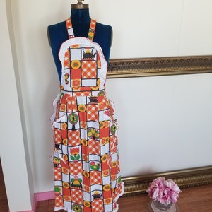 Może przedstawiać: Wzorzysty fartuch w kratę w kolorze pomarańczowym i białym z falbanką i kieszenią z motywem czajnika i kwiatów.