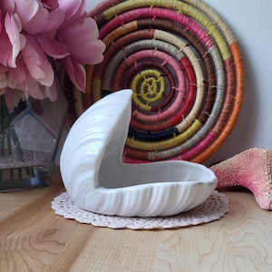 May include: A white ceramic seashell-shaped dish with a textured surface. The dish sits on a small, round, beige doily. A colourful woven placemat and pink flowers are in the background.