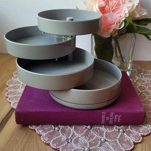 May include: A gray, three-tiered rotating organizer. The organizer is sitting on a purple book and a decorative pink and white beaded doily. A vase of pink flowers is in the background.