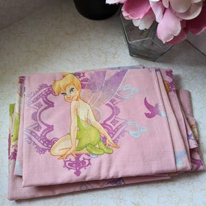 Peut inclure: Ensemble de literie rose avec un motif de personnage de la fée Clochette. La literie présente un motif floral violet et blanc et un personnage de la fée Clochette avec des ailes vertes et une robe verte.