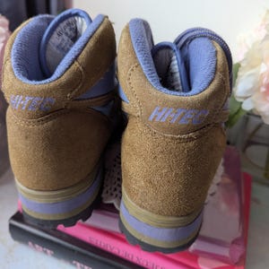 May include: A pair of brown suede hiking boots with blue accents and the word "Hi-Tec" on the side. The boots are sitting on top of a stack of books.