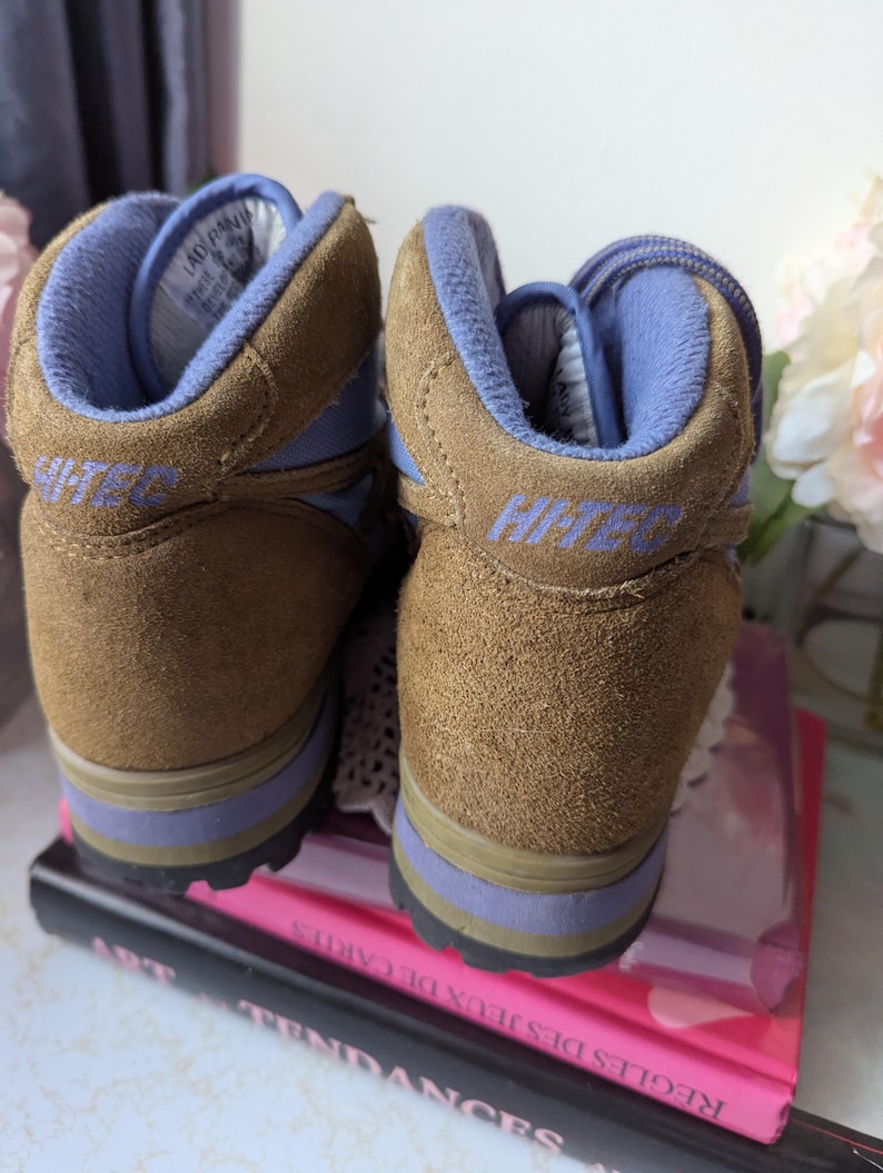 May include: A pair of brown suede hiking boots with blue accents and the word "Hi-Tec" on the side. The boots are sitting on top of a stack of books.