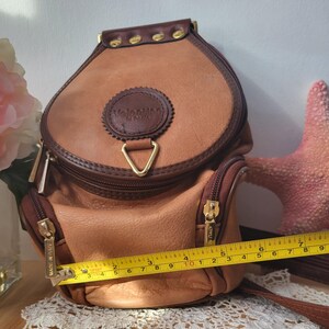 May include: A tan leather Valentino di cuoio backpack with gold accents and a triangular zipper pull. The bag has two zippered compartments and a label that reads "MADE IN ITALY".
