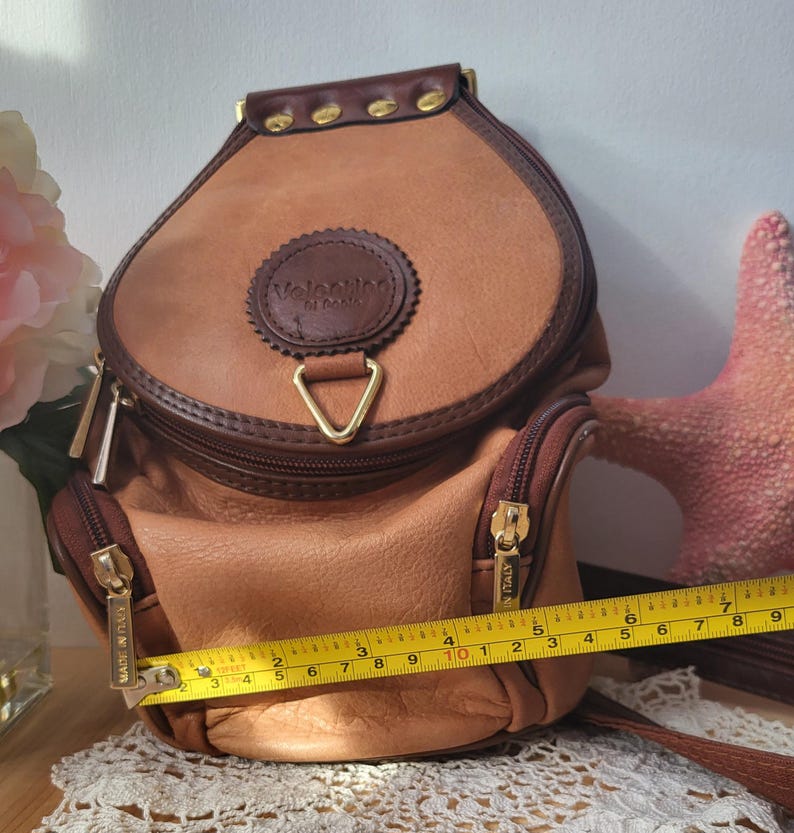 May include: A tan leather Valentino di cuoio backpack with gold accents and a triangular zipper pull. The bag has two zippered compartments and a label that reads "MADE IN ITALY".