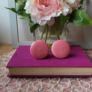 Puede incluir: Dos pomos redondos de color rosa claro descansan sobre un libro morado. Un jarrón de flores rosas y blancas se encuentra detrás de los pomos. El fondo incluye un camino de mesa de cuentas rosas y blancas y una superficie de madera.