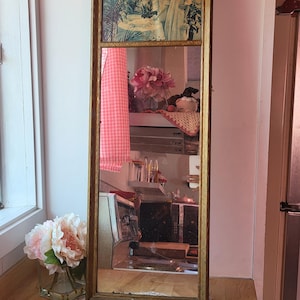 Puede incluir: Un espejo rectangular con marco dorado. La sección superior presenta una impresión vintage con el texto "TROIS MARIAGES REUSSIS". El espejo refleja una habitación con ventana, flores y un reloj.