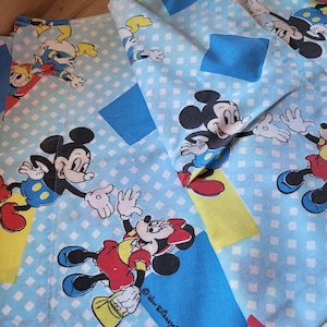 Könnte beinhalten: Hellblauer Stoff mit Mickey- und Minnie-Mouse-Illustrationen. Der Stoff hat ein sich wiederholendes Muster von Mickey und Minnie in verschiedenen Posen, mit blauen, gelben und roten Akzenten. Der Hintergrund ist hellblau mit weißen Rauten.