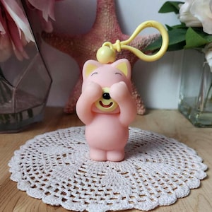 May include: A pink and yellow plastic keychain with a cartoon animal covering its eyes with its paws. The animal is smiling and has a yellow nose and mouth.