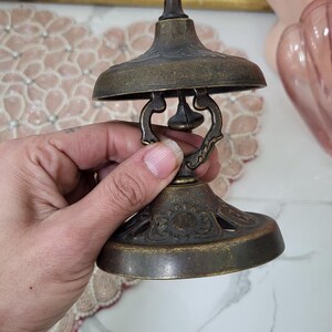 Puede incluir: Campana de lat&oacute;n antiguo con un dise&ntilde;o decorativo. La campana tiene una base acampanada, una parte superior en forma de c&uacute;pula y un peque&ntilde;o asa. La campana se sostiene en una mano y el fondo est&aacute; borroso.