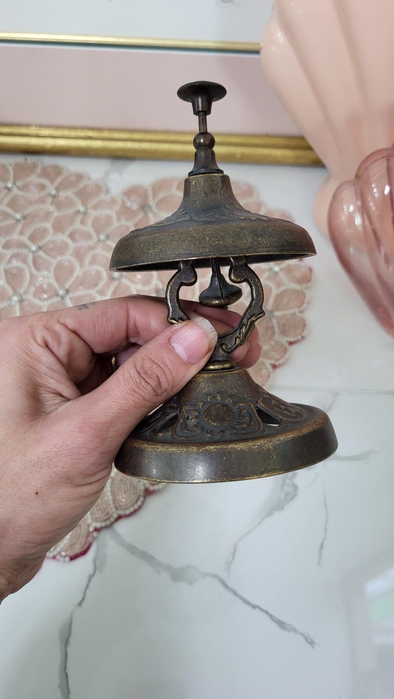 Puede incluir: Campana de lat&oacute;n antiguo con un dise&ntilde;o decorativo. La campana tiene una base acampanada, una parte superior en forma de c&uacute;pula y un peque&ntilde;o asa. La campana se sostiene en una mano y el fondo est&aacute; borroso.