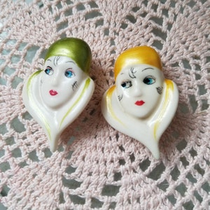 K&ouml;nnte beinhalten: Zwei Keramikfiguren von Frauenk&ouml;pfen mit bemalten Gesichtsz&uuml;gen. Die Figuren sind wei&szlig; mit goldenen und gr&uuml;nen Akzenten. Die Gesichter haben bemalte blaue Augen, rote Lippen und schwarze Augenbrauen. Die Figuren stehen auf einer rosa geh&auml;kelten Oberfl&auml;che.