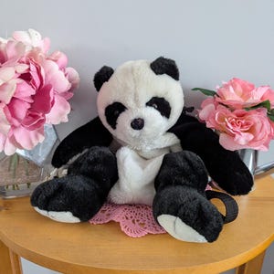 May include: A black and white plush panda bear sitting on a wooden table with a pink crocheted doily. The panda has a black nose and black eyes.