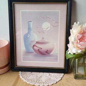 Op de afbeelding: Een ingelijste print van een stilleven met een blauwe vaas en een roze pot met deksel. De achtergrond is een zachte roze en paarse gradi&euml;nt met een tak met witte bloesems en een volle maan.