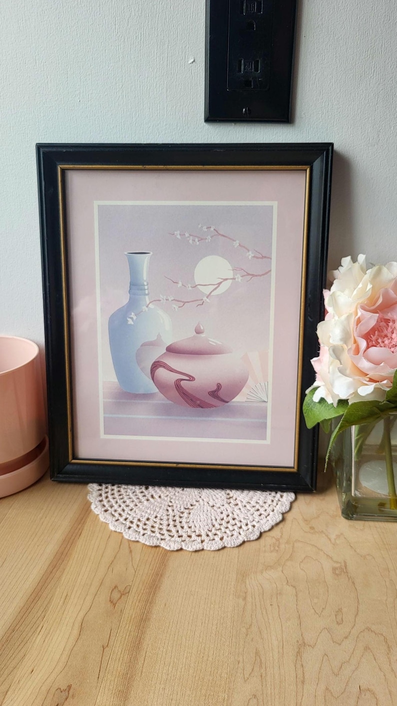 Op de afbeelding: Een ingelijste print van een stilleven met een blauwe vaas en een roze pot met deksel. De achtergrond is een zachte roze en paarse gradi&euml;nt met een tak met witte bloesems en een volle maan.