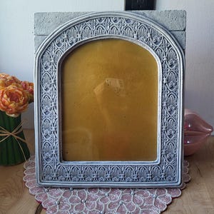 May include: A grey, arched picture frame with ornate detailing. The frame has a light grey finish and a decorative pattern around the edges. It sits on a pink beaded doily. A vase of orange flowers and a pink lip-shaped object are in the background.