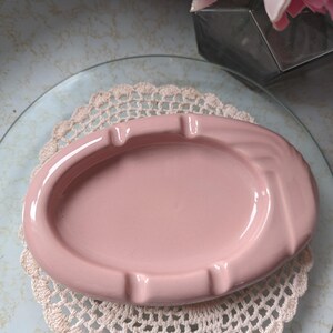 May include: A pink ceramic ashtray with a rounded oval shape. The ashtray has a smooth, glossy finish and a slight indentation in the center.