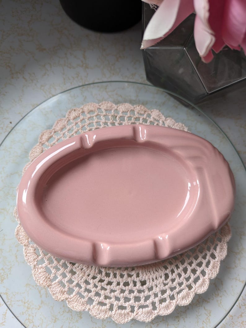 May include: A pink ceramic ashtray with a rounded oval shape. The ashtray has a smooth, glossy finish and a slight indentation in the center.