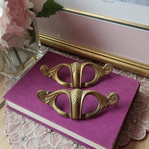 May include: Two antique brass drawer pulls with a decorative design, resting on a purple book. The pulls have a curved shape with ornate detailing. A framed picture and flowers are in the background.