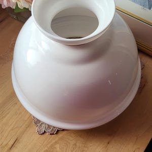 May include: A white, glossy, vintage-style lampshade with a rounded base and a flared top. The lampshade is sitting on a decorative doily. The background includes a framed picture and a bouquet of pink flowers.