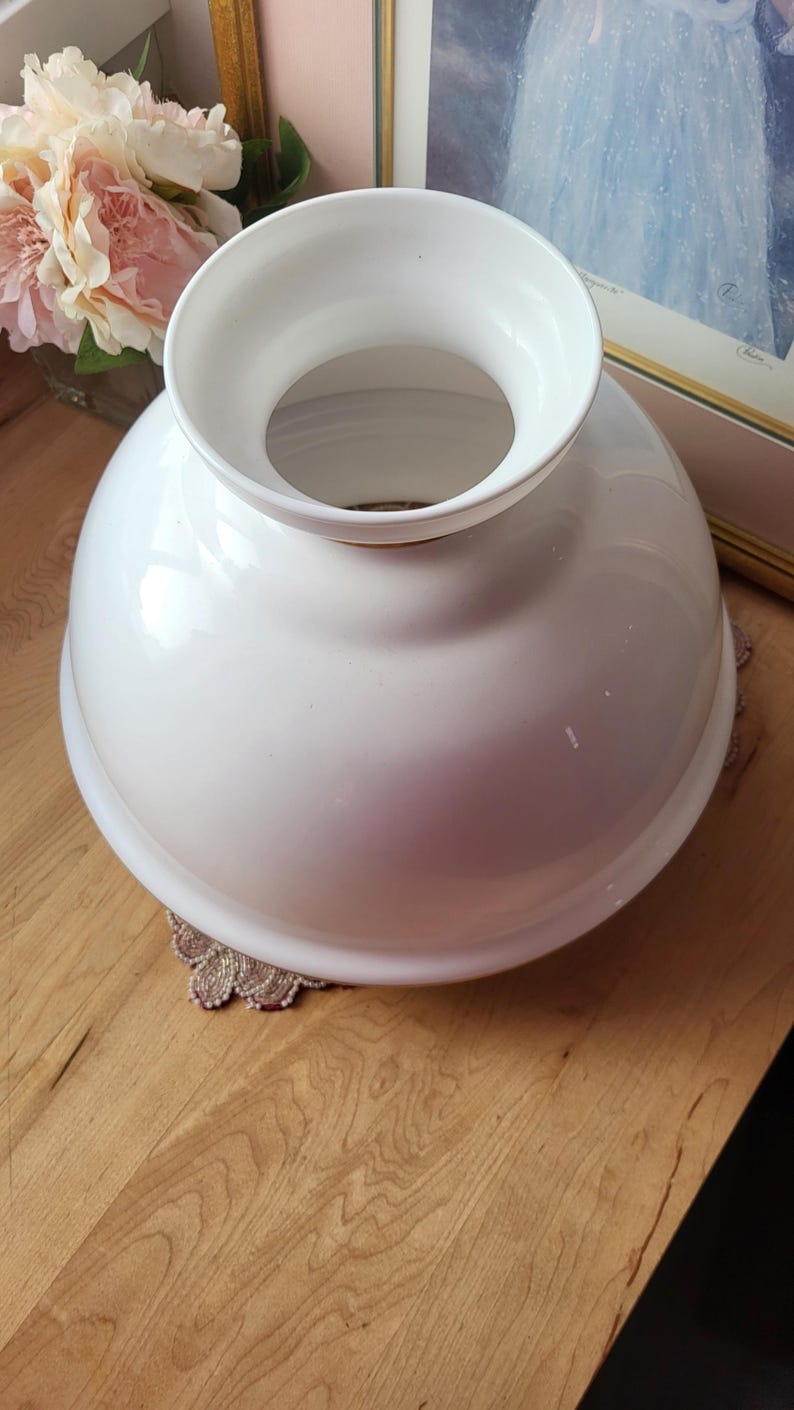 May include: A white, glossy, vintage-style lampshade with a rounded base and a flared top. The lampshade is sitting on a decorative doily. The background includes a framed picture and a bouquet of pink flowers.