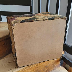 May include: A rectangular storage ottoman with a floral tapestry fabric top and a brown wood base. The ottoman is sitting on wooden stairs.