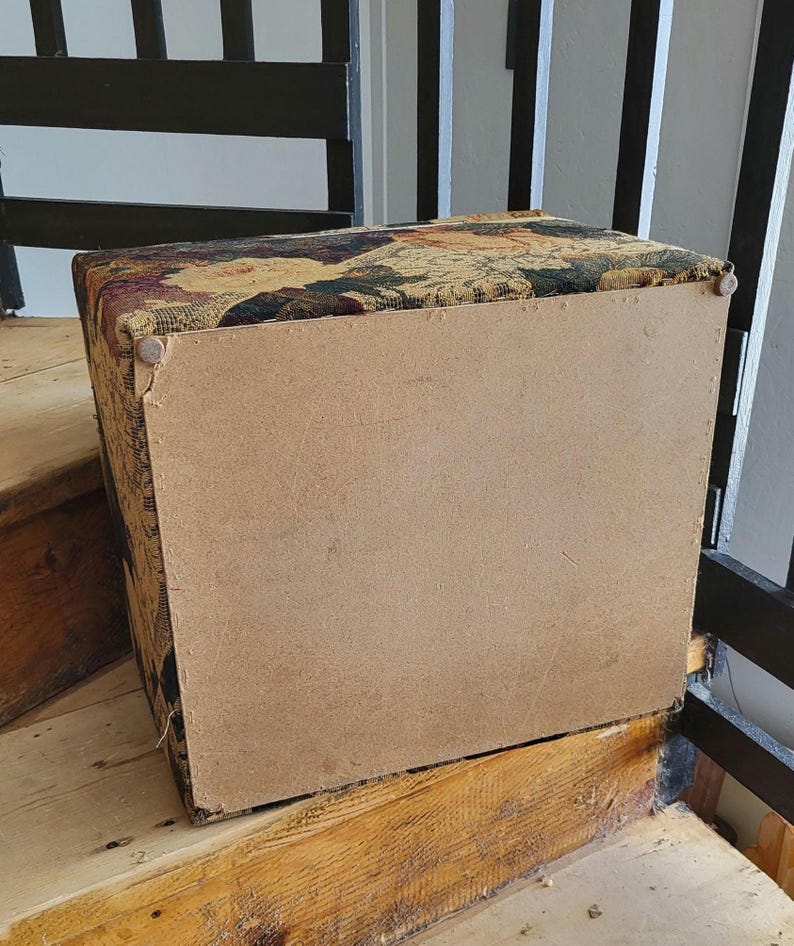 May include: A rectangular storage ottoman with a floral tapestry fabric top and a brown wood base. The ottoman is sitting on wooden stairs.