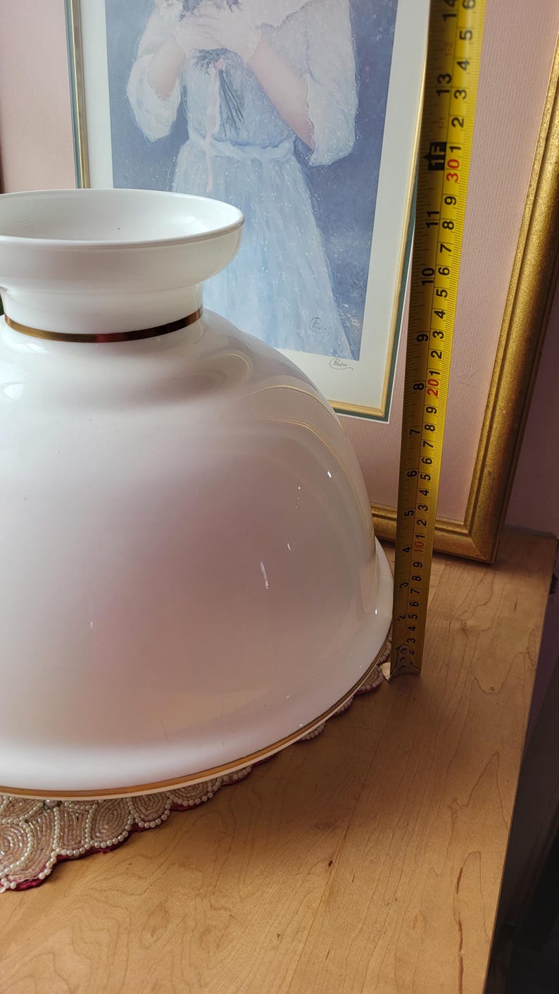 May include: A large, white ceramic bowl with a gold rim, approximately 10 inches in diameter. The bowl has a wide, shallow shape and sits on a decorative surface. A framed picture is in the background.