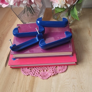 May include: Four blue plastic drawer pulls with silver screws, stacked on top of two books with pink covers. The books are on a wooden surface with a pink crocheted doily.