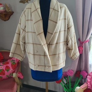 May include: Cream-colored, double-breasted jacket with a brown plaid pattern. The jacket has a wide lapel collar and is displayed on a mannequin with a blue top. The jacket has two buttons and two pockets.