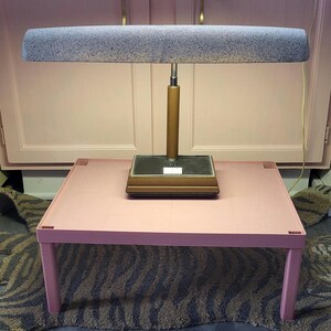 Puede incluir: Una lámpara de escritorio vintage con una pantalla gris moteada y un vástago color latón se encuentra sobre una mesa rosa. La lámpara es rectangular con una base marrón. La mesa también es rosa y tiene un diseño sencillo. El fondo incluye armarios rosas.