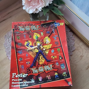 Peut inclure: Une boîte de puzzle rouge Yu-Gi-Oh! avec des illustrations de personnages et le logo du jeu. La boîte comprend également le texte "Porter Puzzle Rompecabezas Casse-tête".
