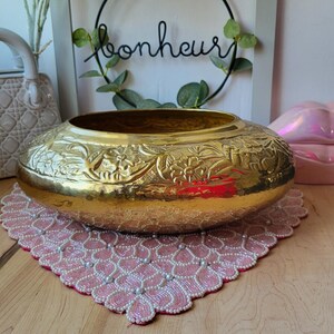 May include: A large, gold-coloured, embossed brass bowl with floral designs. The bowl is sitting on a pink beaded placemat on a wooden surface.