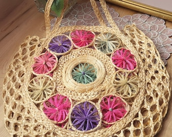 Può includere: Borsa tote rotonda in paglia intrecciata con accenti floreali colorati. La borsa presenta un design a griglia e un motivo circolare di fiori rosa, viola e verdi. Ha due manici intrecciati.