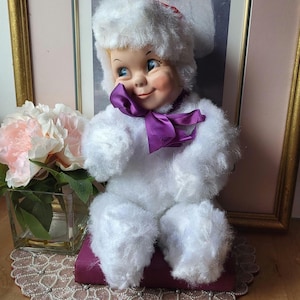 Può includere: Una bambola vintage con un viso sorridente, occhi blu e un abito bianco e peloso, compreso un cappello e un fiocco. La bambola è seduta su un cuscino viola, con un'immagine incorniciata sullo sfondo. Un mazzo di fiori rosa è in un vaso di vetro.