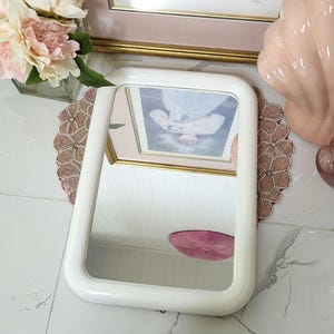 May include: A white rectangular mirror with rounded corners. The mirror reflects a framed picture and a pink object. The mirror is placed on a decorative doily. Other decorative items include a framed picture, flowers, and a pink seashell.