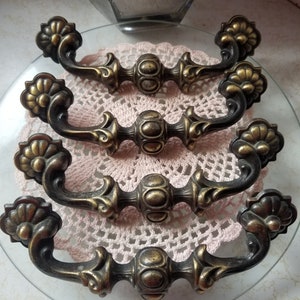 Może przedstawiać: Zestaw pięciu antycznych mosiężnych gałek do szafek z ozdobnymi wzorami kwiatowymi i zaokrąglonym środkiem. Gałki są ułożone na białej serwetce.