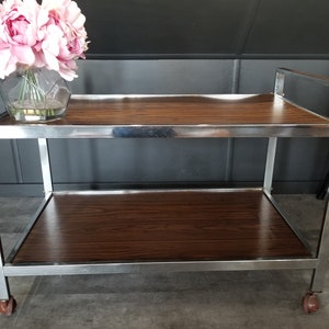 May include: A chrome serving cart with two wooden shelves. The cart has a modern design and is perfect for entertaining guests.