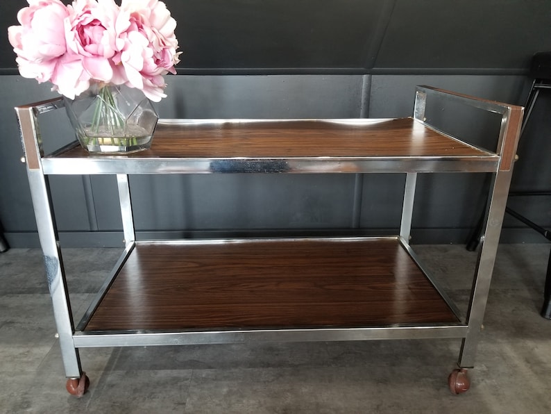 May include: A chrome serving cart with two wooden shelves. The cart has a modern design and is perfect for entertaining guests.