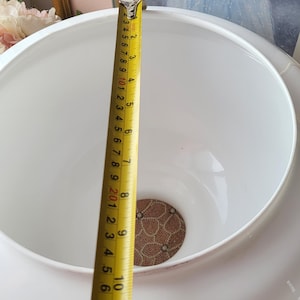 May include: A white ceramic bowl with a wide rim, measuring approximately 11 inches in height. A yellow tape measure is extended inside the bowl, showing the measurement. The bowl has a decorative, patterned base.
