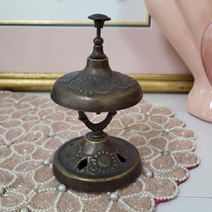 Puede incluir: Campana de lat&oacute;n antiguo con detalles florales. La campana tiene una parte superior en forma de c&uacute;pula y una base circular, conectadas por un asa curva. La campana est&aacute; colocada sobre un mantel individual rosa con cuentas. Una pintura enmarcada est&aacute; en el fondo.