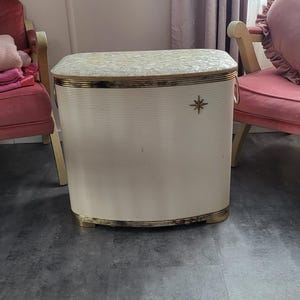 Könnte beinhalten: Vintage cremefarbener Wäschekorb mit gemustertem Deckel und goldfarbenen Zierleisten. Der Korb hat ein Sternmuster und Metallgriffe. Die vertikale Rippenstruktur verleiht dieser Aufbewahrungslösung einen klassischen Touch.