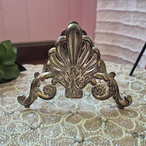 May include: A silver-toned metal napkin holder featuring an ornate, symmetrical design with a central fan-shaped motif, scrolling flourishes, and small circular floral accents at the base, supported by curved feet on each side.