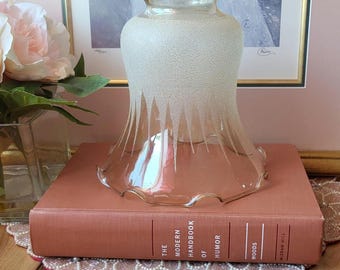 May include: A clear glass lampshade with a textured, frosted finish and a scalloped edge. The lampshade is sitting on a vintage book titled "The Modern Handbook of Humor". The background includes a framed picture and a pink floral arrangement.