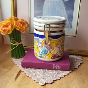 May include: A white ceramic jar with a lid, featuring a colourful abstract design in yellow, blue, and purple. The jar has a gold-coloured metal clasp and trim. It sits on a purple book and a decorative doily.