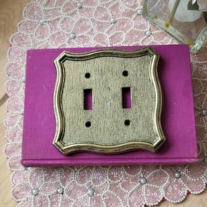 Puede incluir: Una cubierta de interruptor doble dorada con una superficie texturizada y un marco decorativo. Descansa sobre un libro morado. La placa del interruptor tiene dos aberturas rectangulares para los interruptores y dos aberturas circulares para los tornillos. El fondo incluye un mantel individual rosa con cuentas.