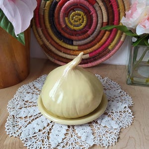 May include: A yellow ceramic butter dish shaped like a garlic bulb. The dish is sitting on a white crocheted doily on a wooden surface.