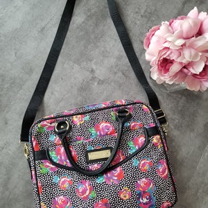 May include: Black laptop bag with a floral print featuring pink, red, and green roses on a white polka dot background. The bag has a black adjustable shoulder strap and two black handles. The bag has a gold metal zipper and a gold metal logo that says "Juicy Couture".