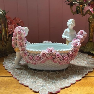 Puede incluir: Un centro de mesa de cerámica blanca con borde festoneado, adornado con rosas rosas y figuras de querubines. El cuenco está sostenido por dos querubines, cada uno sosteniendo una guirnalda de rosas. La pieza está sobre un tapete de encaje.