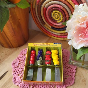 May include: A set of four stainless steel spreaders with colourful fruit-shaped handles. The handles are red apple, purple grapes, red strawberries, and yellow lemon. The box is yellow with the text "Spread It On" and "Gourmet Spreader Set".