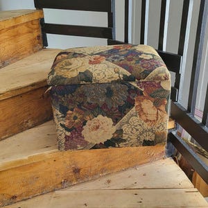 May include: A floral patterned storage ottoman with a square top. The ottoman is sitting on wooden stairs.